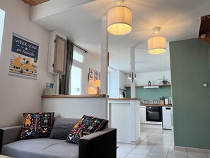 Un salon moderne avec un canapé, des coussins colorés, une cuisine en arrière-plan et des luminaires suspendus.