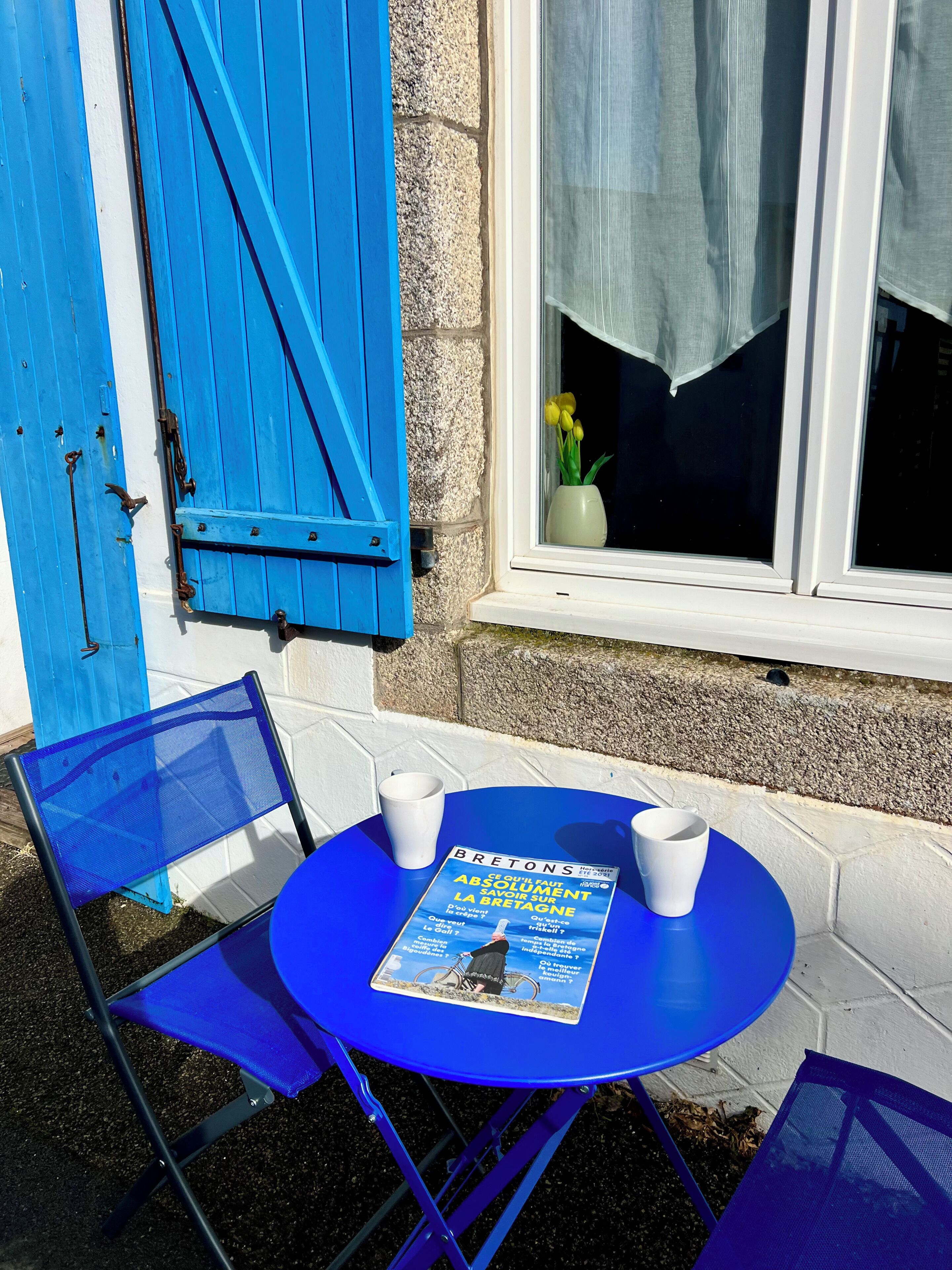 Une table avec un magazine, deux chaises et une fenêtre donnant sur un immeuble, ainsi qu’un vase rempli de fleurs.