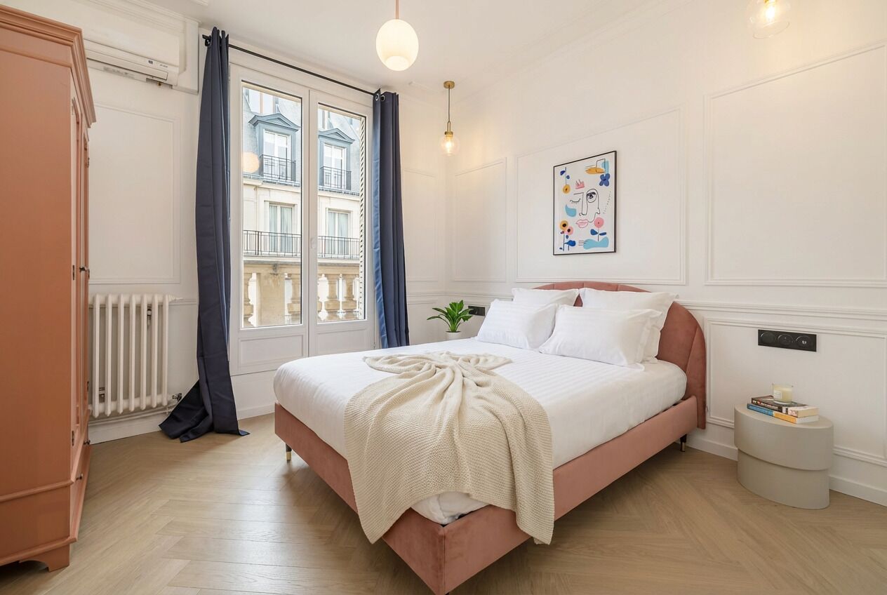 A bedroom with a bed, a radiator, a window with curtains, a bedside table, and a wall-mounted light fixture.