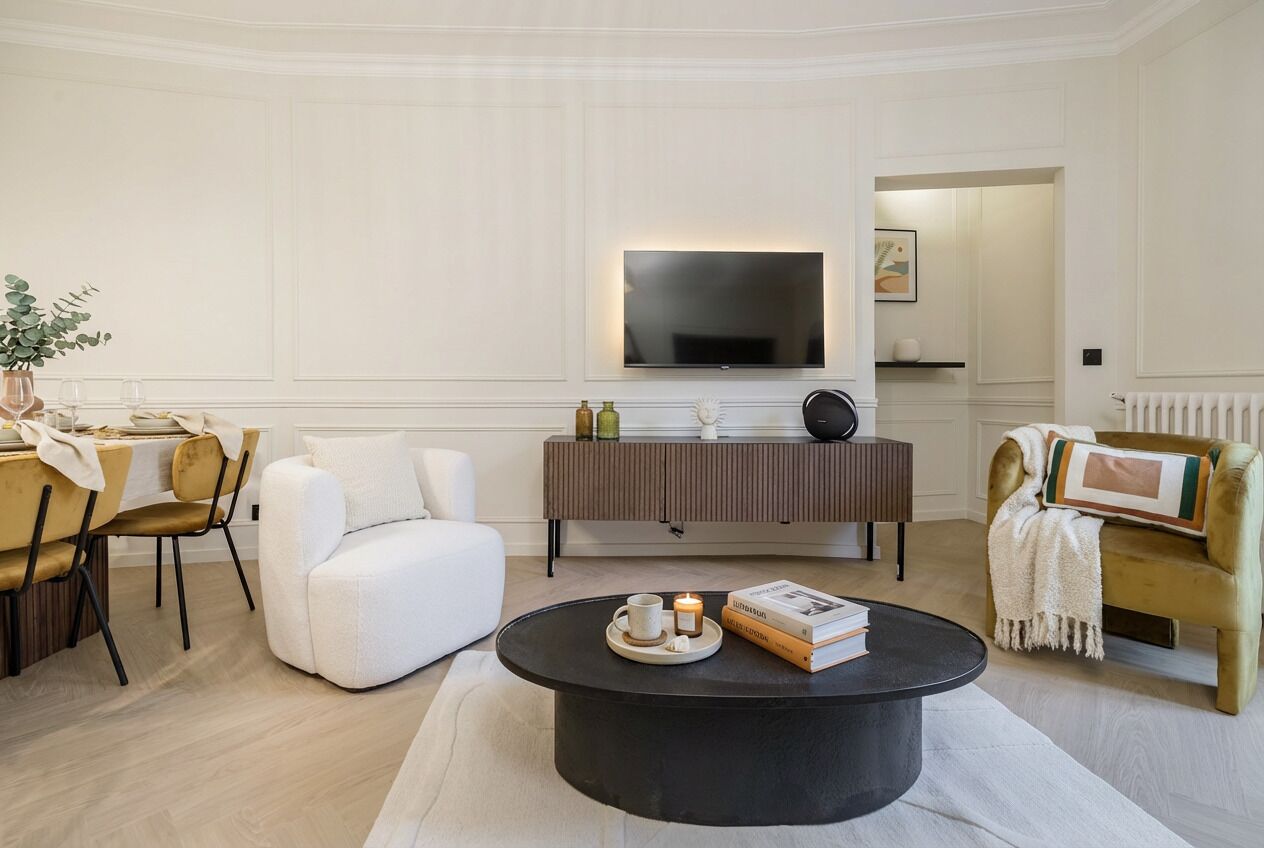 Uma sala de estar moderna com uma televisão de tela plana, uma mesa de centro redonda com livros e uma vela, um sofá e uma área de jantar com cadeiras e uma mesa.
