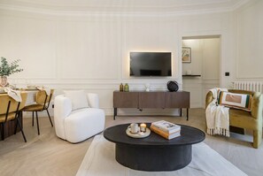 Uma sala de estar moderna com uma televisão de tela plana, uma mesa de centro redonda com livros e uma vela, um sofá e uma área de jantar com cadeiras e uma mesa.