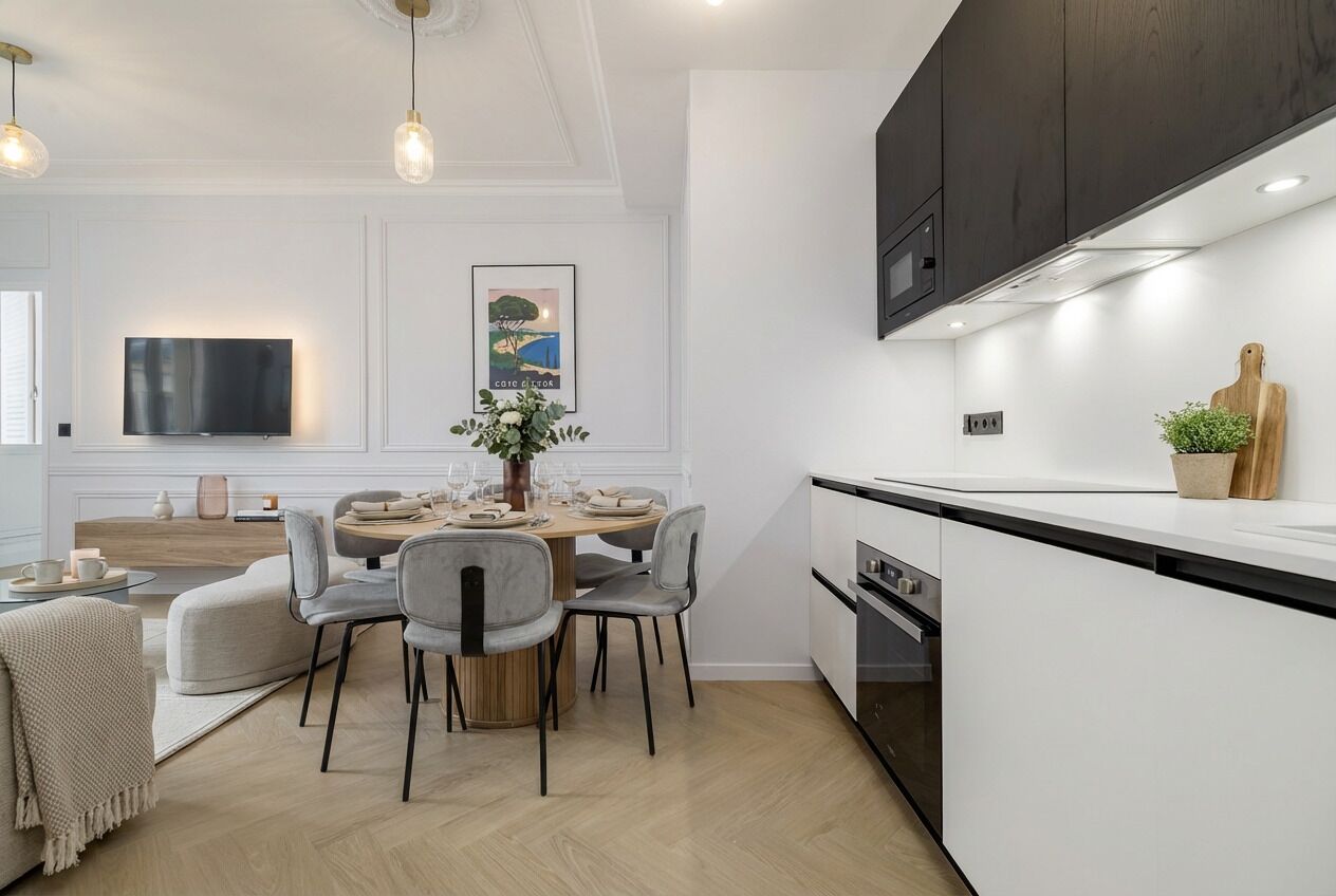 Een moderne keuken met witte kasten, een eethoek met een ronde tafel en stoelen, en een flatscreen televisie aan de muur.