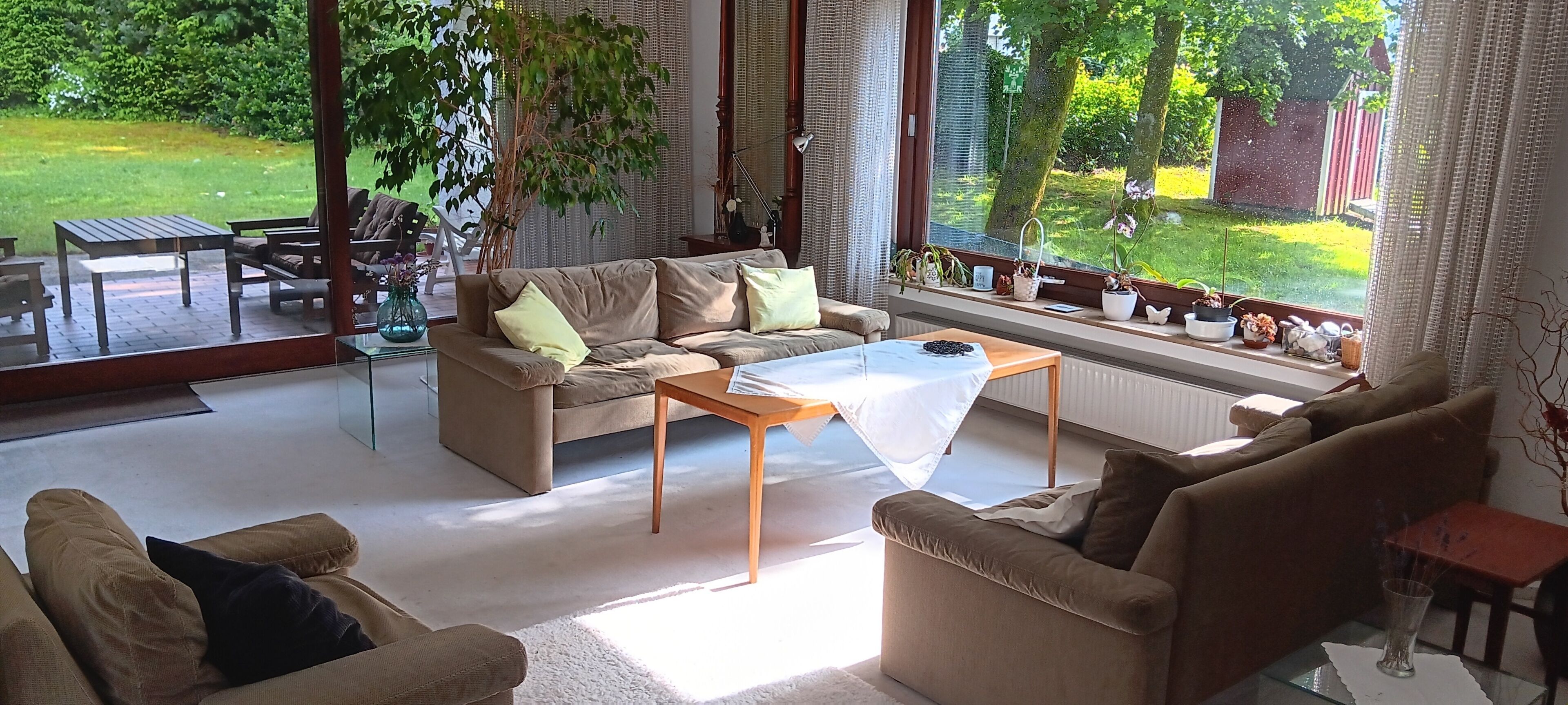 Ein modernes Wohnzimmer mit einer Couch, Sesseln, einem Couchtisch und Blick auf einen Garten durch große Fenster.
