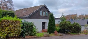 Ein eingeschossiges Haus mit dunklem Dach und weißen Außenwänden, umgeben von verschiedenen Sträuchern und wolkenlosem Himmel.