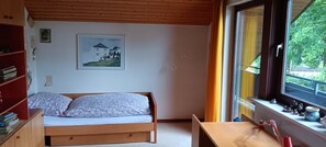 Une chambre à coucher avec un lit, une bibliothèque, un tableau encadré au mur et une fenêtre donnant sur un paysage verdoyant.
