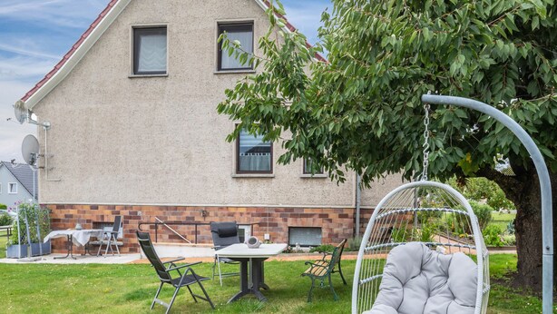 Ett hus med en grund av tegel, ett stort träd och utemöbler, inklusive en gungstol och ett bord.
