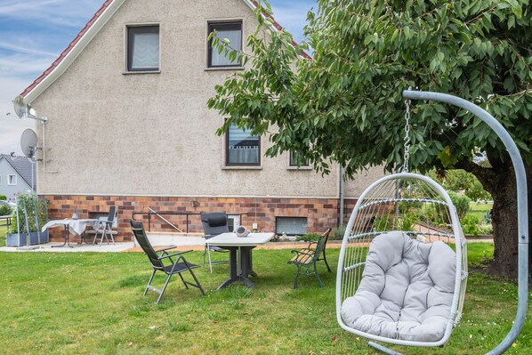 Una casa con base de ladrillo, un árbol grande y mobiliario exterior que incluye una silla colgante y una mesa.