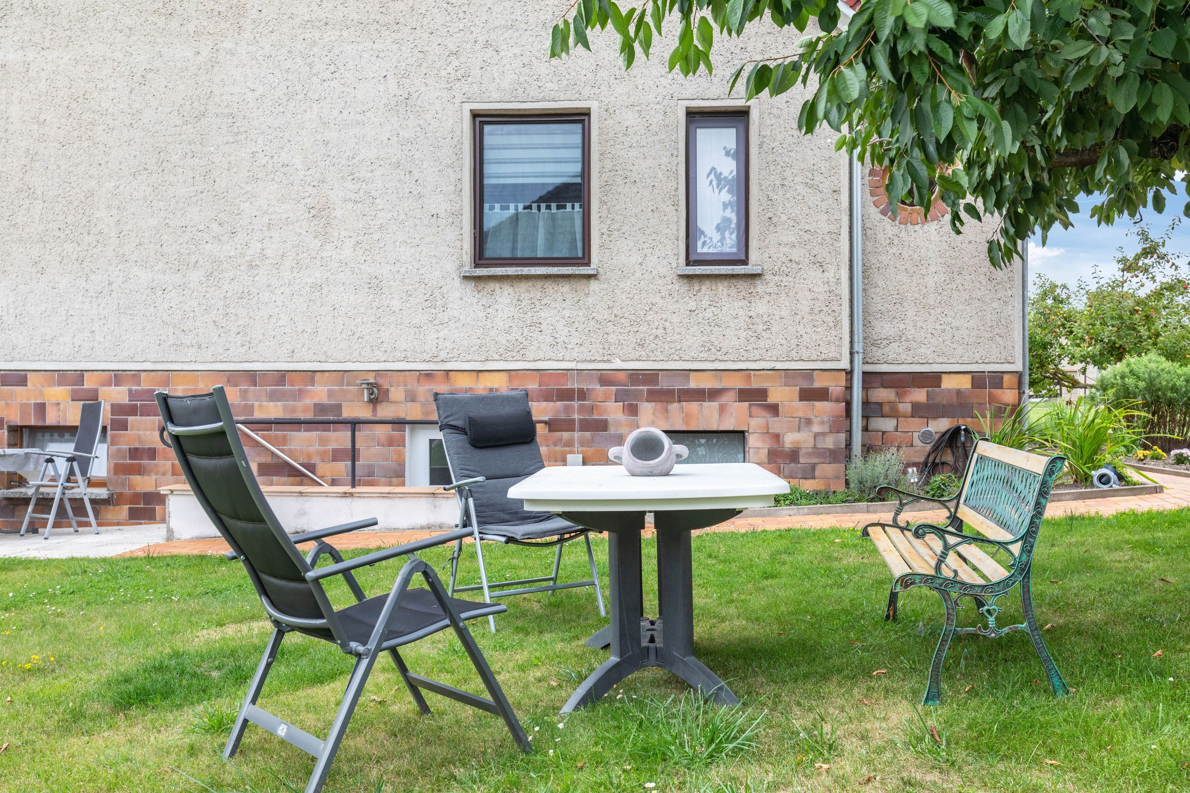 Ein Sitzbereich im Freien mit Tisch und Stühlen, einer Bank und einer Ziegelwand mit Fenstern.