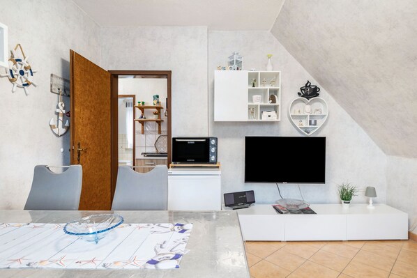 A modern living room with a flat-screen TV, a white entertainment unit, and a dining area with a table set for two.