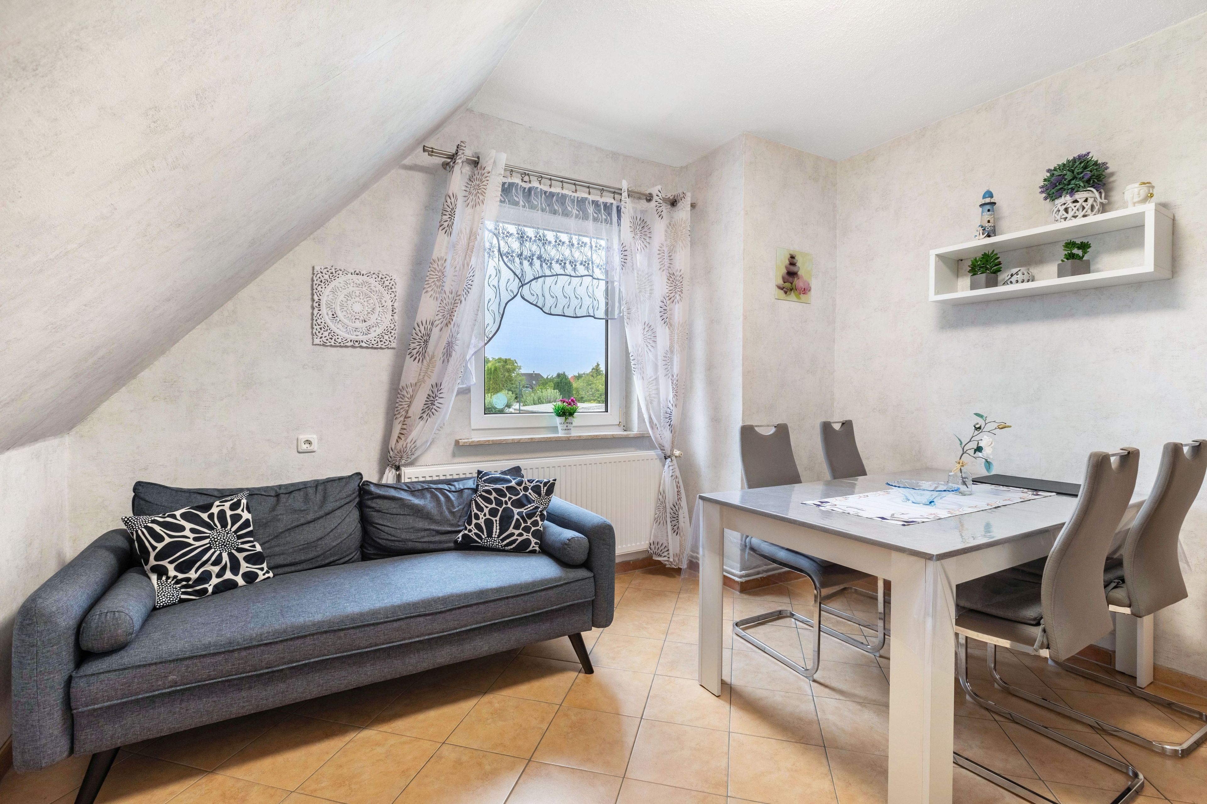 Ein gemütliches Zimmer mit grauem Sofa, einem Esstisch mit Stühlen, einem Fenster mit Vorhängen und einem Regal mit Dekoration.
