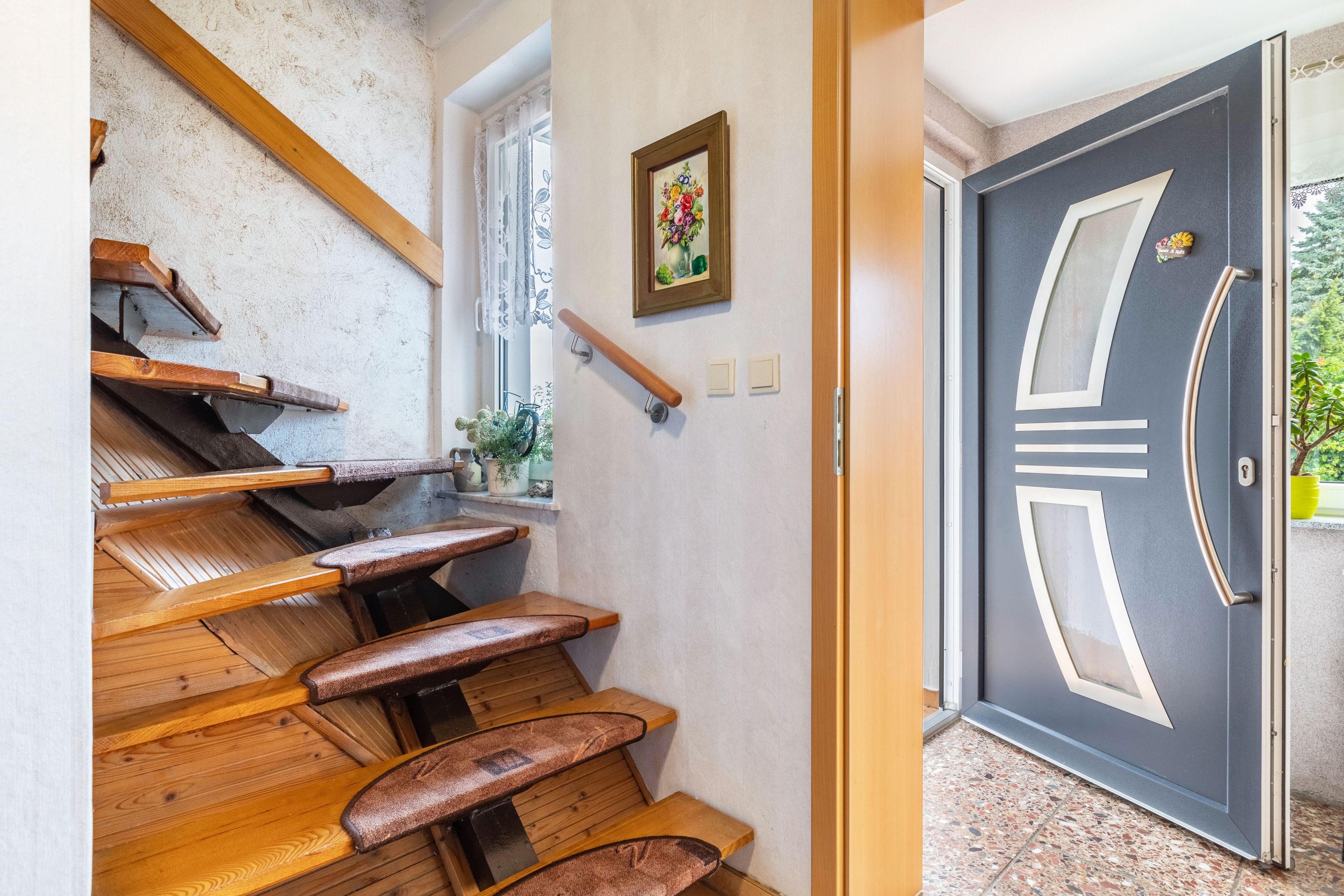Eine Holztreppe mit Handlauf, eine Tür mit dekorativem Griff und ein Fenster mit Vorhang.