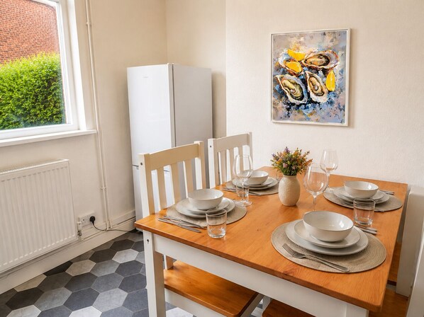 A dining area with a wooden table set for four, white chairs, and a white refrigerator in the background.