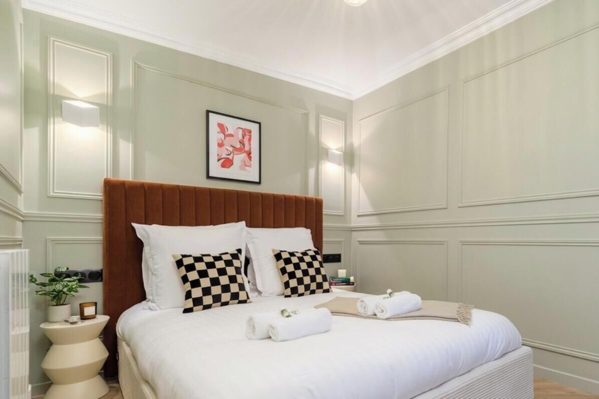 A neatly made bed with white linens and checkered pillows, a bedside table with a plant, and a wall-mounted light fixture.