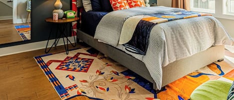 Un dormitorio con cama, mesita de noche, lámpara, ventana con cortinas y una alfombra colorida.