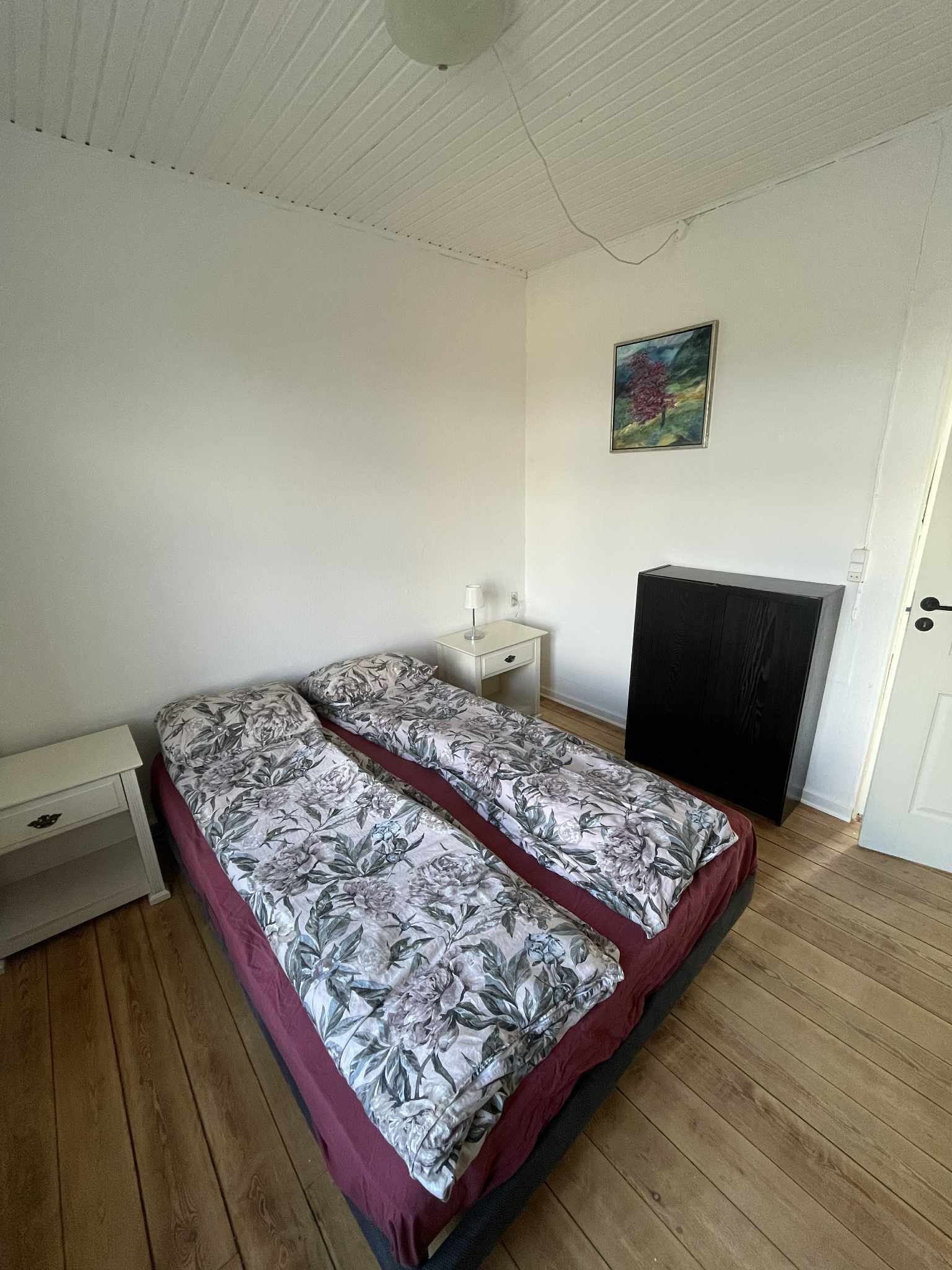 A room with two beds, a white nightstand, a desk, a chair, a painting on the wall, and a door.