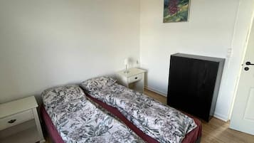 A room with two beds, a white nightstand, a desk, a chair, a painting on the wall, and a door.