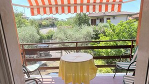 A balcony with a yellow table and two chairs, a striped awning, and a view of greenery and buildings.