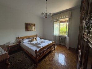 A bedroom with a wooden bed, a nightstand, a bedside lamp, a window with curtains, and a radiator. - Two-bedroom apartment on the first floor, just a short walk from downtown (Bardolino)