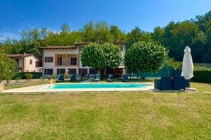 A two-story house with a swimming pool, outdoor seating, and a large tree in the front yard.