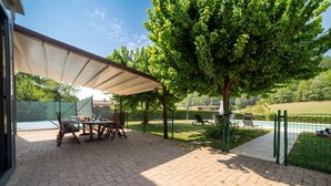 A patio area with a covered table and chairs, a swimming pool in the background, and a tree providing shade.