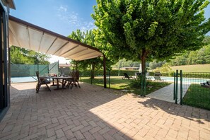 A patio area with a covered table and chairs, a swimming pool in the background, and a tree providing shade.