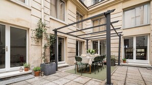 Un patio con una mesa lista para comer, rodeado de plantas en macetas y una pérgola.