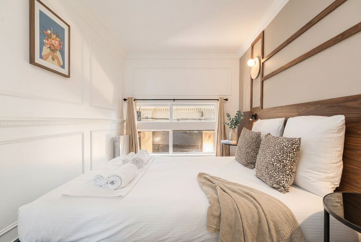 Une chambre d’hôtel avec un grand lit, une fenêtre avec des rideaux, un tableau au mur et une table de chevet avec une lampe.