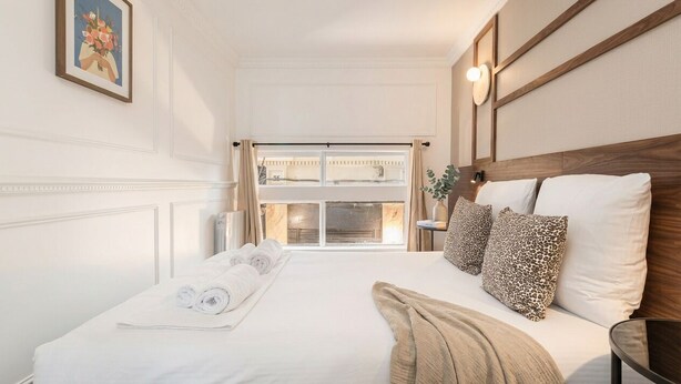 Habitación de hotel con una cama grande, una ventana con cortinas, un cuadro en la pared y una mesita de noche con una lámpara.
