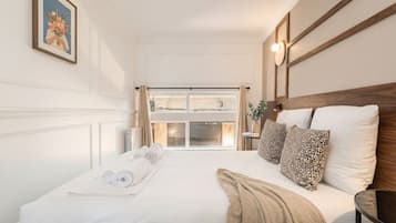 A hotel room with a large bed, a window with curtains, a painting on the wall, and a bedside table with a lamp.