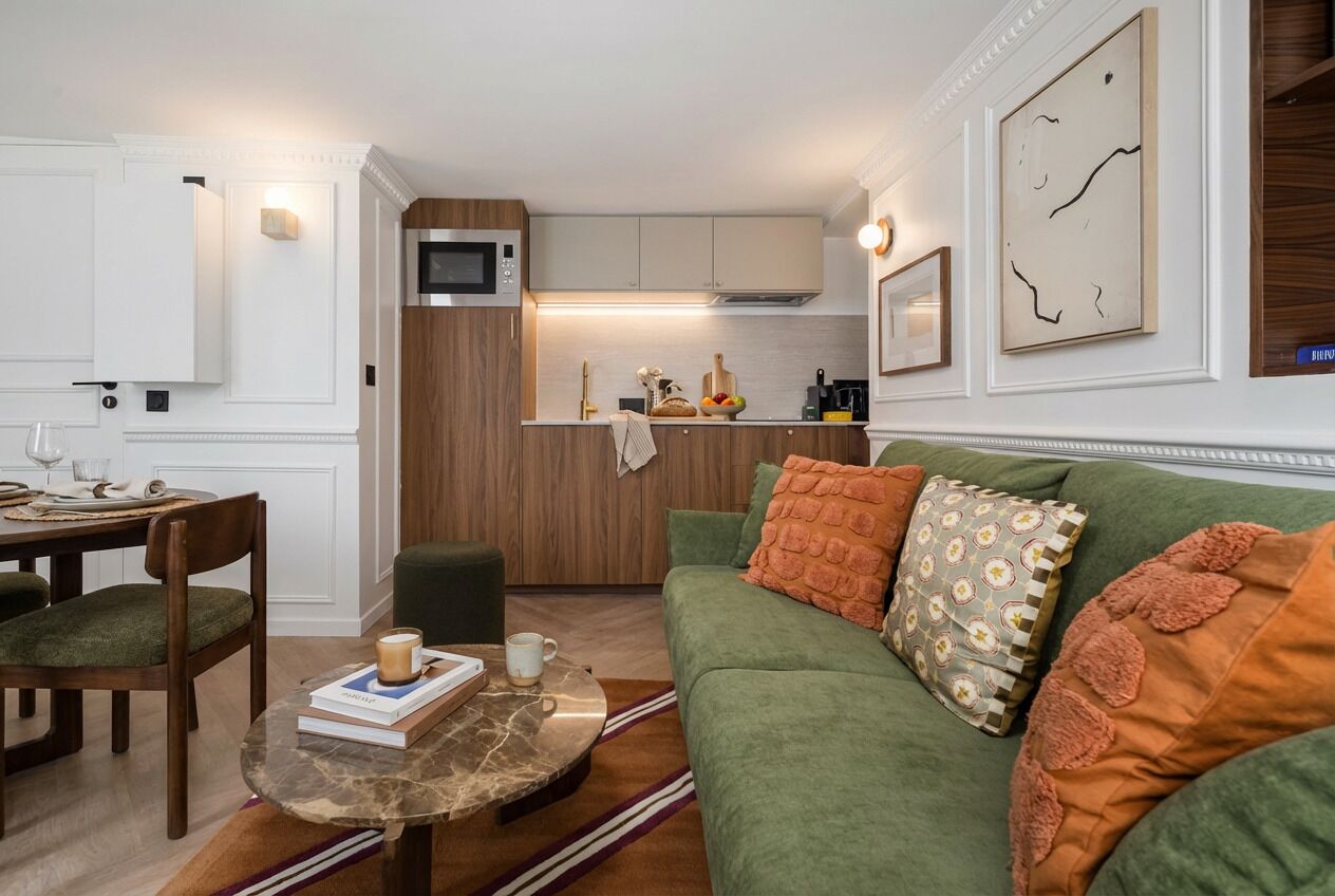 Un espacio habitable compacto con comedor, un sofá verde con cojines estampados, una pequeña cocina con microondas y una mesa de comedor con sillas.