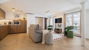 Una sala de estar moderna con un sofá gris, un sillón verde, una zona de cocina con armarios de madera y un ventanal grande.