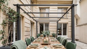 Ett långt matbord i trä, dukat för en måltid med stolar, en pergola ovanför och en krukväxt till vänster.