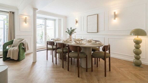 En matplats med ett runt bord dukat för sex personer, omgivet av stolar, med en grön fåtölj och en golvlampa i förgrunden.