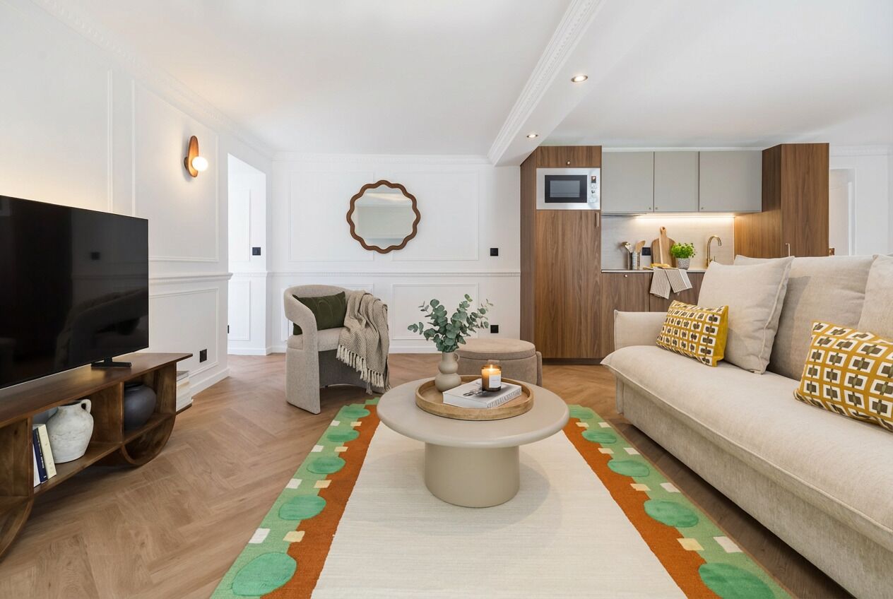 Een moderne woonkamer met een flatscreen televisie, een ronde salontafel, een bank met gestreepte kussens, en een keuken met houten kastjes.