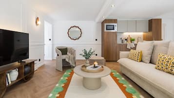 A modern living room with a flat-screen TV, a round coffee table, a sofa with patterned cushions, and a kitchen area with wooden cabinets.