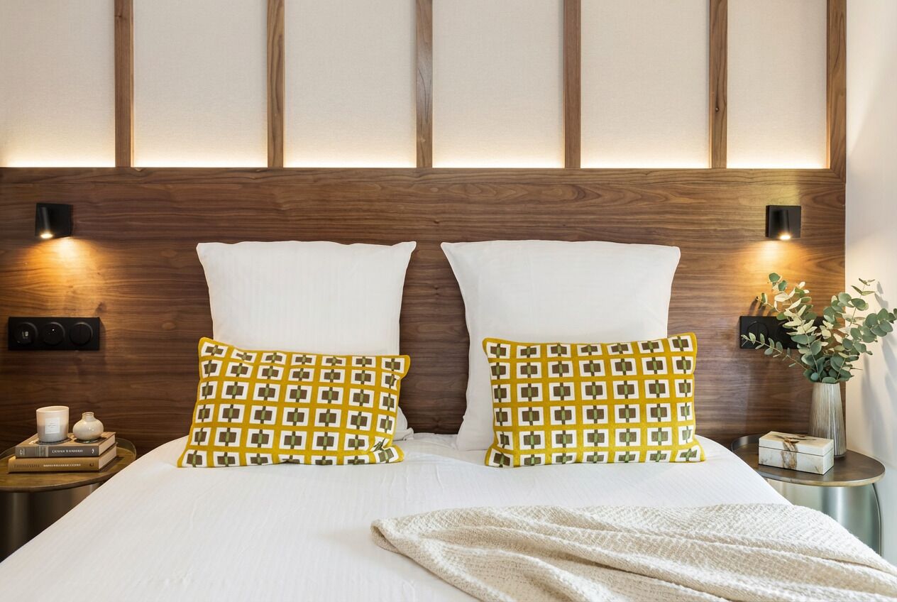 Una cama bien hecha con sábanas blancas y dos almohadas decorativas, una mesita de noche con un libro y una vela, y una lámpara de pared.