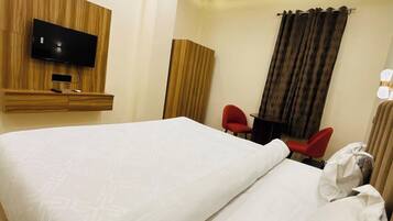 Habitación de hotel con una cama grande, un televisor montado en un panel de madera, un escritorio con dos sillas rojas y una ventana con cortinas.