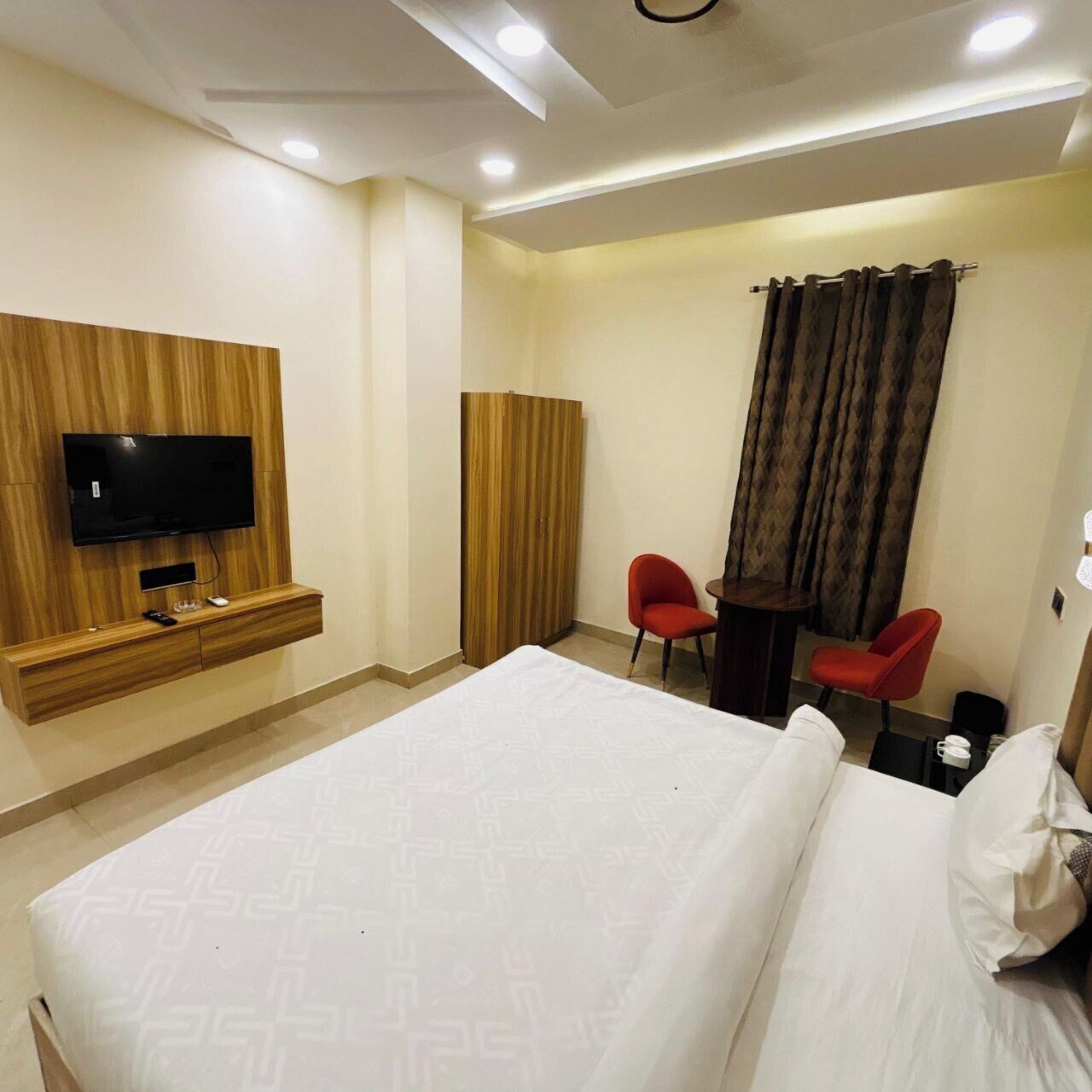 A hotel room with a bed, a TV mounted on a wooden panel wall, a desk with two chairs, and a curtain.