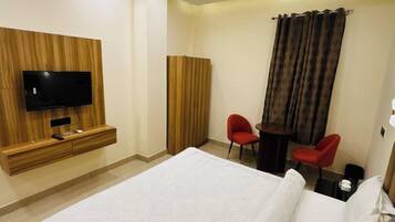 A hotel room with a bed, a TV mounted on a wooden panel wall, a desk with two chairs, and a curtain.