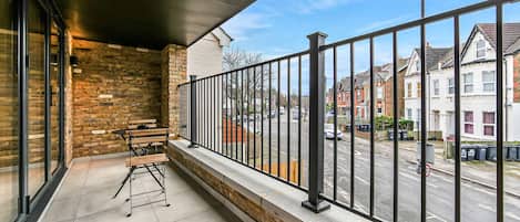 Un balcón con vista a una calle residencial, una baranda de metal y un banco de madera.