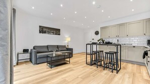 Un salon moderne avec un canapé gris, une table basse et un coin cuisine équipé de tabourets de bar.