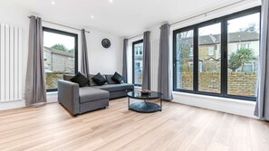 Uma sala de estar moderna com um sofá cinzento, uma mesa de centro redonda e grandes janelas com cortinas.