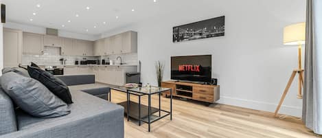Una sala moderna con un sofá gris, una mesa de centro, un televisor mostrando 'NETFLIX' y una cocina al fondo.
