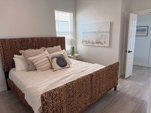Un dormitorio con cabecera trenzada, cama con varios cojines, mesita de noche con lámpara y un cuadro en la pared.