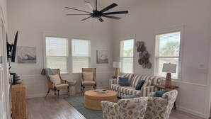 Ein geräumiges Wohnzimmer mit Deckenventilator, zwei Fenstern mit Jalousien, einem gestreiften Sofa, einem runden Couchtisch und einem gemusterten Sessel.
