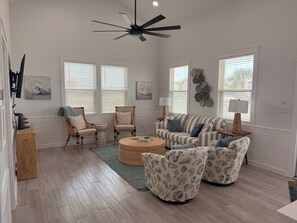Ein geräumiges Wohnzimmer mit Deckenventilator, zwei Fenstern mit Jalousien, einem gestreiften Sofa, einem runden Couchtisch und einem gemusterten Sessel.