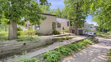 A modern two-story residential building with a flat roof, large windows, and a front yard with a wooden deck and landscaping.