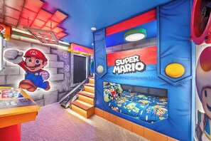 Ein Zimmer im Super-Mario-Design, mit einem Bett mit Mario-Bettwäsche, einer Wand mit einem Mario-Wandgemälde und einer Spielecke mit Figuren von Mario und Luigi.