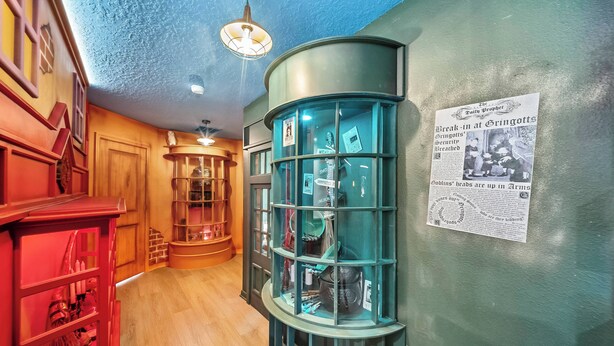 Um corredor com uma vitrine curva exibindo diversos objetos, um armário vermelho com objetos decorativos e uma parede com um recorte de jornal.