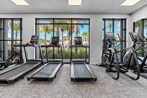 Un gimnasio con cintas de correr y elípticas, amplias ventanas que dan a vistas de palmeras y un cielo despejado.